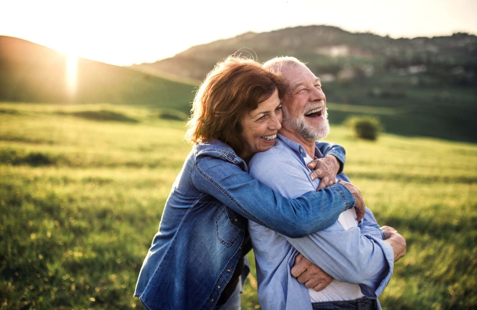 People Happy About Hearing Aid Financing Woman holds a man in a field from behind. He is leaning into her and laughing with his arms crossed. Her arms are draped over his shoulders. She is smiling and the sun is going down on the hillside behind them.